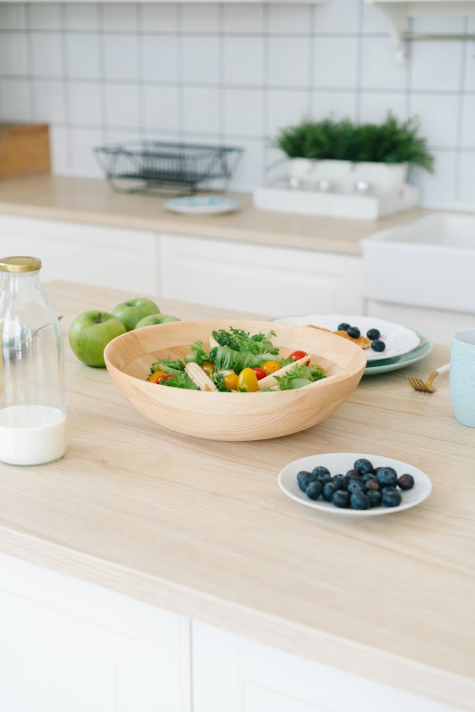A fresh and healthy kitchen display with salad, blueberries, milk, and apples on a wooden counter.
